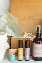 balanced home apothecary best sellers bundle that includes a white basket, calm hanging car diffuser, joyful rollerball, sleep rollerball, and hormones magnesium spray, with a brown box, mint tissue paper, and beige nest.