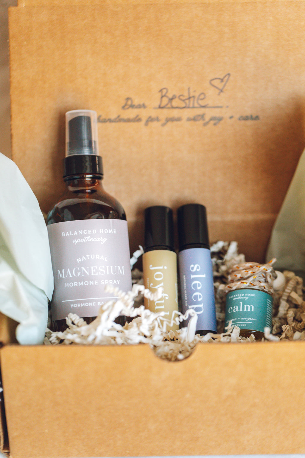 Set of skincare products in a brown gift box with personalized text. balanced home apothecary best sellers bundle that includes a white basket, calm hanging car diffuser, joyful rollerball, sleep rollerball, and hormones magnesium spray, with a brown box, mint tissue paper, and beige nest.