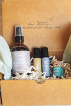 Set of skincare products in a brown gift box with personalized text. balanced home apothecary best sellers bundle that includes a white basket, calm hanging car diffuser, joyful rollerball, sleep rollerball, and hormones magnesium spray, with a brown box, mint tissue paper, and beige nest.