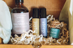 Collection of natural health products including a bottle labeled 'Balanced Home' and small containers labeled 'joyful', 'sleep', and 'calm' in a box with shredded paper. balanced home apothecary best sellers bundle that includes a white basket, calm hanging car diffuser, joyful rollerball, sleep rollerball, and hormones magnesium spray, with a brown box, mint tissue paper, and beige nest.