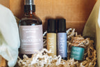 Set of natural health products including bottles and sprays in a box with shredded paper. balanced home apothecary best sellers bundle that includes a white basket, calm hanging car diffuser, joyful rollerball, sleep rollerball, and hormones magnesium spray, with a brown box, mint tissue paper, and beige nest.